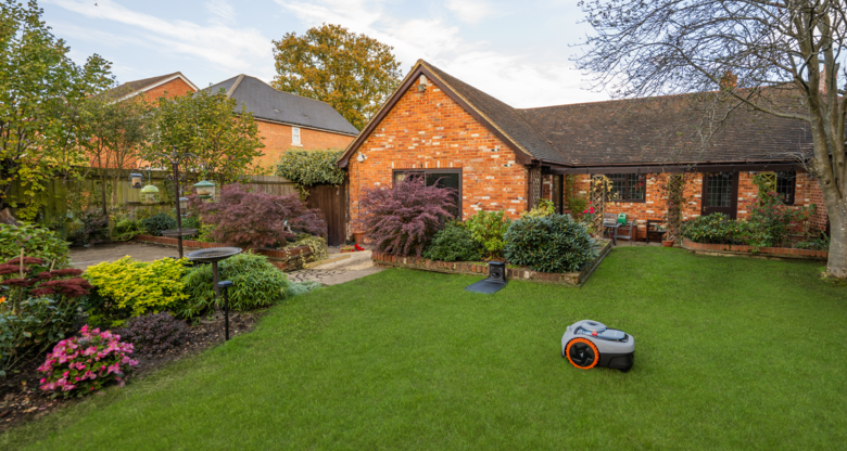 Herbstlich bepflanzter Garten mit Vogeltränke, Büschen in kräftigen Farben und Mähroboter vor charmantem Landhaus.
