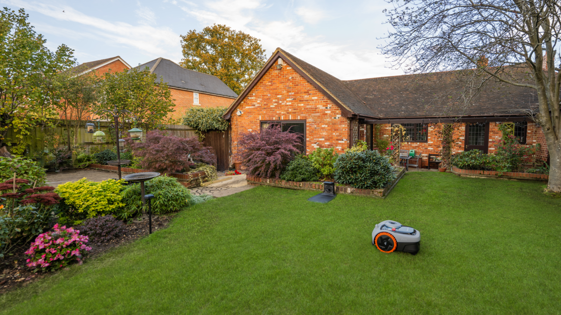 Herbstlich bepflanzter Garten mit Vogeltränke, Büschen in kräftigen Farben und Mähroboter vor charmantem Landhaus.